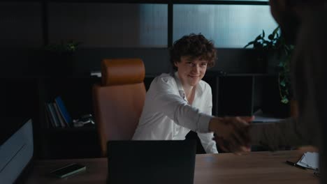 Happy-young-guy-with-curly-hair-in-a-white-shirt-shaking-hands-with-an-experienced-colleague-during-the-start-of-an-interview-in-the-office