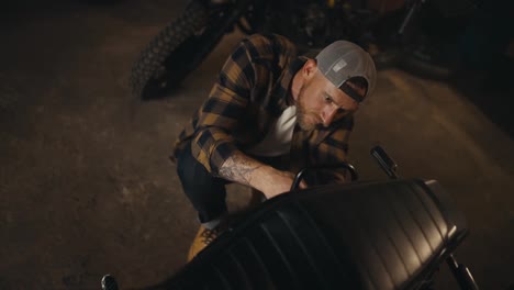 Top-view-of-a-guy-mechanic-in-a-plaid-shirt-and-cap-repairing-a-motorcycle-in-his-workshop-studio