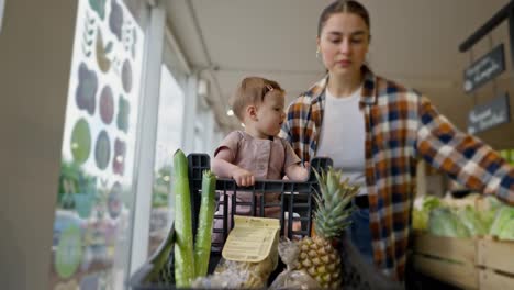 Interested-little-girl-baby-rides-in-a-cart-and-helps-her-mom-shopping-in-a-supermarket.-A-brunette-woman-and-her-little-daughter-choose-goods-during-their-shopping-in-a-supermarket