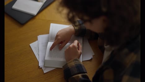 Over-the-shoulder,-a-concentrated-girl-student-with-curly-hair-reads-a-book-while-sitting-at-a-table-in-the-library