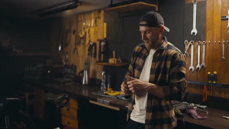 Happy-guy-mechanic-drinks-soda-from-a-glass-bottle-after-finishing-his-working-day-in-a-cozy-workshop-studio