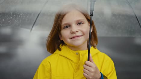 Portrait-of-a-happy-blonde-teenage-girl-in-a-yellow-jacket-holding-a-transparent-umbrella-on-a-walk-after-the-rain