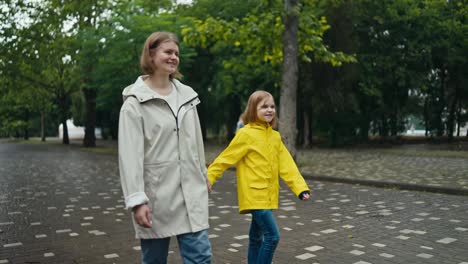 A-happy-blonde-woman-in-a-white-jacket-walks-hand-in-hand-with-her-teenage-daughter-in-a-yellow-jacket-walking-in-the-park-after-the-rain