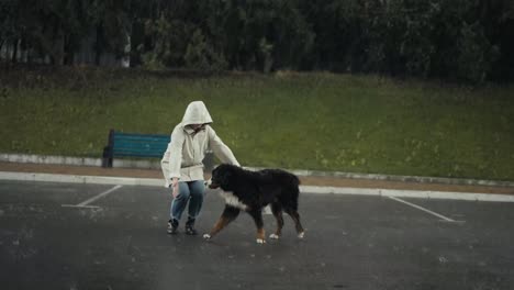 Mujer-Feliz-Con-Una-Chaqueta-Blanca-Acariciando-A-Su-Perro-Grande-Mientras-Camina-Bajo-Una-Fuerte-Lluvia-En-El-Parque