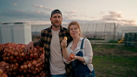 Portrait-of-a-Happy-farmer-guy-with-his-girlfriend-eating-peaches-near-bags-of-onions-after-a-successful-harvest-season-on-the-farm