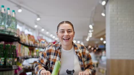 Porträt-Eines-Glücklichen-Brünetten-Mädchens-In-Einem-Karierten-Hemd,-Das-Rennt-Und-Dann-Auf-Einen-Einkaufswagen-Springt-Und-In-Einen-Supermarkt-Fährt