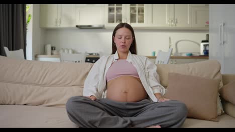 A-pregnant-brunette-woman-in-gray-pants-and-a-white-shirt-sits-on-the-sofa-and-meditates-at-home-while-expecting-a-baby