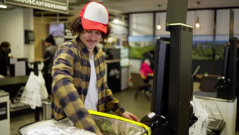 Confident-food-delivery-guy-in-a-plaid-shirt-punches-goods-at-the-checkout-and-then-puts-them-in-his-yellow-bag-for-food-delivery-in-the-supermarket