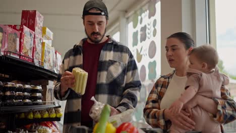 Confident-brunette-man-with-a-beard-in-a-cap-together-with-his-wife-and-small-child-choose-goods-during-their-family-shopping-in-a-supermarket