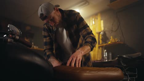 Confident-male-mechanic-in-a-checkered-shirt-is-repairing-a-motorcycle-and-selecting-the-necessary-spare-parts-in-his-workshop-studio