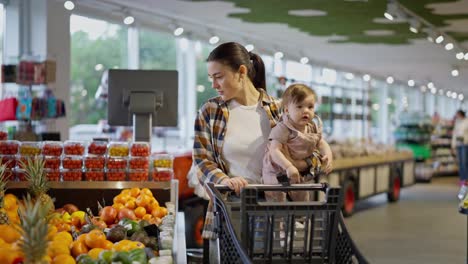 Una-Niña-Morena-Con-Una-Camisa-A-Cuadros-Sostiene-A-Su-Pequeña-Hija-En-Brazos-Y-Empuja-Un-Carrito-En-Un-Supermercado-Mientras-Hace-Compras.