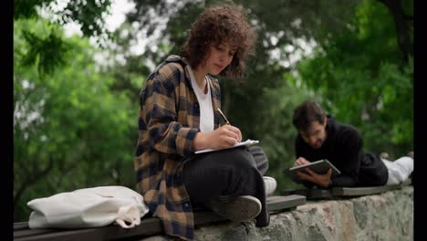 A-brunette-student-girl-with-curly-hair-in-a-brown-shirt-sits-on-a-bench-and-makes-notes-in-a-notebook-near-her-fellow-guy-in-the-park