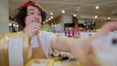 A-brunette-guy-with-curly-hair-in-a-cap-chews-a-bun-and-takes-inventory-while-rearranging-goods-on-the-shelves-in-a-supermarket