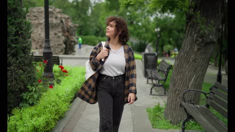 Una-Chica-Morena-Feliz-Con-Cabello-Rizado-Con-Una-Camisa-A-Cuadros-Y-Una-Camiseta-Blanca-Con-Una-Mochila-Blanca-Camina-Por-El-Parque-Después-De-Sus-Clases-En-La-Universidad.