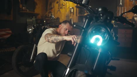 Focused-guy-mechanic-with-a-tattoo-inspects-and-repairs-a-motorcycle-in-his-garage-workshop