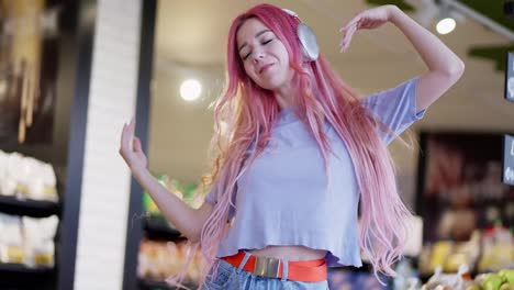 Happy-girl-with-pink-hair-in-a-purple-T-shirt-wearing-white-headphones-listens-to-music-and-dances-in-a-supermarket