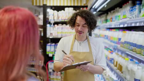 Über-Der-Schulter-Hält-Ein-Glücklicher-Kerl-Mit-Lockigem-Haar-Ein-Tablet-In-Den-Händen-Und-Macht-Eine-Inventur,-Während-Er-Mit-Kunden-In-Der-Milchprodukteabteilung-Eines-Supermarkts-Kommuniziert