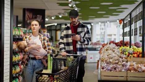 A-man-in-a-plaid-shirt-looks-at-the-grocery-list-on-his-phone-with-his-wife-and-small-infant-while-shopping-in-a-supermarket