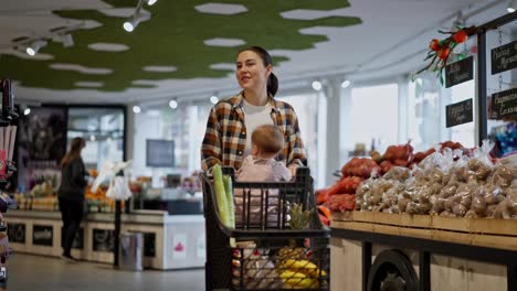 A-happy-brunette-girl-in-a-plaid-shirt-pushes-a-cart-along-with-her-little-child-and-groceries-during-her-shopping-in-a-supermarket