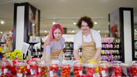 Una-Chica-De-Cabello-Rosa-Junto-Con-Su-Asistente,-Un-Chico-Moreno-Con-Un-Delantal-Amarillo,-Están-Haciendo-Un-Inventario-Y-Reorganizando-Las-Frutas-En-El-Departamento-De-Cítricos-De-Un-Supermercado.