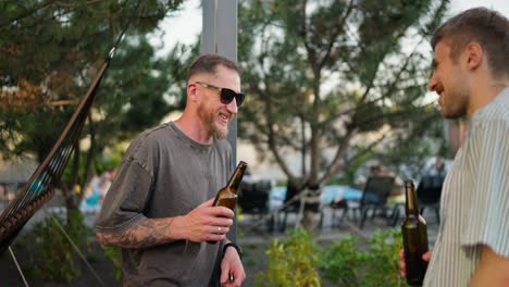 Over-his-shoulder-a-confident-guy-in-sunglasses-with-a-beard-talks-and-drinks-beer-from-a-brown-bottle-with-his-friend-in-the-courtyard-of-a-country-house