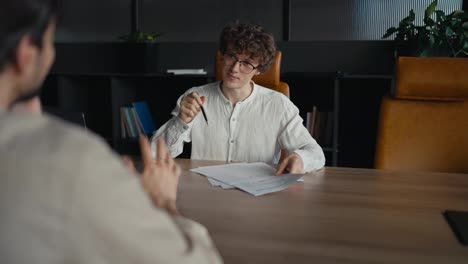 Over-his-shoulder-a-young-guy-with-curly-hair-wearing-glasses-and-a-white-T-shirt-is-communicating-with-a-more-experienced-colleague-about-specific-documents-and-important-papers-at-the-table-in-the-office