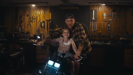 Portrait-of-a-happy-male-mechanic-with-a-beard-together-with-his-beloved-daughter-in-a-workshop-studio