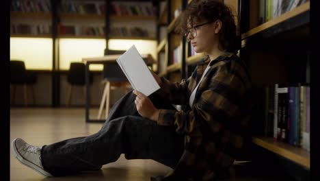 Confident-girl-with-curly-hair-and-glasses-sits-near-shelves-with-books-and-reads-one-in-the-library