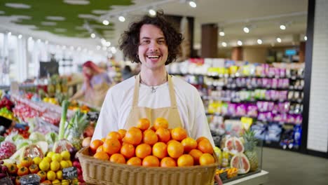 Porträt-Eines-Glücklichen-Brünetten-Kerls-Mit-Lockigem-Haar,-Der-In-Seinen-Händen-Eine-Kiste-Mit-Vielen-Orangefarbenen-Früchten-In-Einem-Supermarkt-Hält