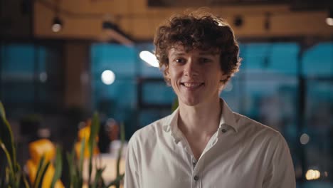 Portrait-of-a-happy-young-guy-with-curly-hair-in-a-white-shirt-who-is-posing-and-smiling-in-the-evening-office