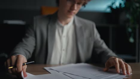 Close-up-a-confident-young-guy-with-curly-hair-in-a-gray-jacket-examines-documents-and-signs-while-sitting-at-a-table-in-the-office