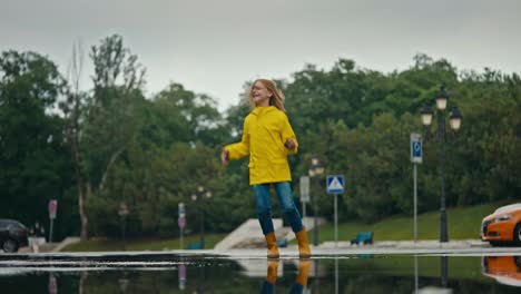 Fröhliches-Teenager-Mädchen-In-Gelber-Jacke-Und-Orangefarbenen-Gummistiefeln-Springt-Nach-Dem-Regen-In-Eine-Pfütze-Im-Park