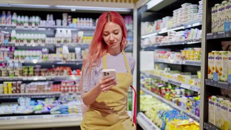 A-girl-with-pink-hair-in-a-yellow-apron-scrolls-through-social-networks-during-a-break-at-work-among-dairy-products-in-a-supermarket