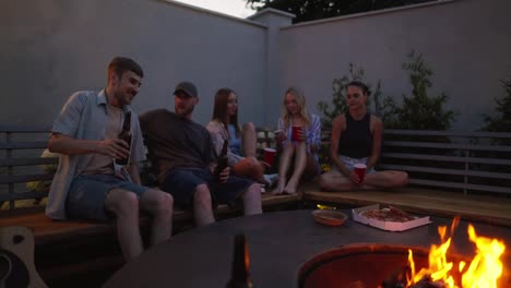 A-group-of-friends-are-sitting-near-a-fire,-chatting-and-having-a-happy-time-while-relaxing-in-the-courtyard-of-a-country-house