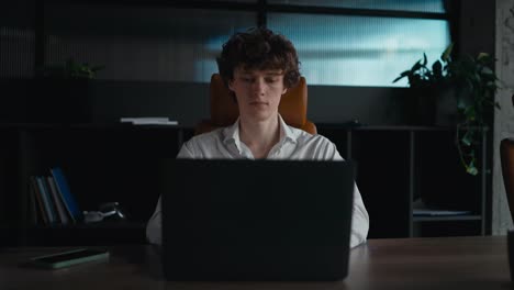 Portrait-of-a-confident-guy-with-curly-hair-in-a-white-shirt-who-works-at-a-gray-laptop-in-the-office-and-looks-at-the-camera-without-a-smile-on-his-face