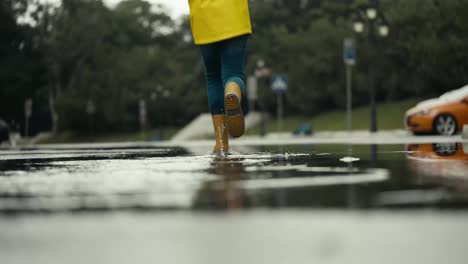 Nahaufnahme-Eines-Glücklichen-Teenager-Mädchens-In-Einer-Gelben-Jacke-Und-Gummistiefeln,-Das-Nach-Regen-Im-Park-Durch-Eine-Pfütze-Rennt-Und-Wasser-An-Die-Seiten-Spritzt