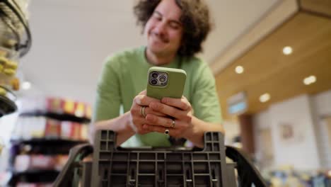 Curly-brunette-guy-uses-his-green-smartphone-while-texting-while-shopping-in-a-supermarket