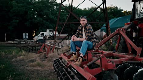 Portrait-of-a-happy-farmer-guy-in-a-cap-and-plaid-shirt-who-is-sitting-on-a-combine-harvester-and-posing-near-a-field-on-a-farm