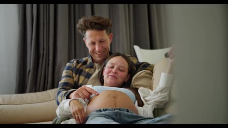 Happy-middle-aged-man-in-a-plaid-shirt-stroking-the-belly-of-his-pregnant-wife-while-sitting-on-the-sofa-at-home-while-relaxing