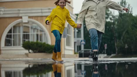 Una-Niña-Feliz-Con-Una-Chaqueta-Amarilla-Y-Botas-De-Goma-Camina-Por-Un-Charco-Con-Su-Madre-Y-Salpica-Agua-A-Los-Lados-Del-Parque-Después-De-La-Lluvia.