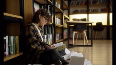 Vergrößern-Sie-Die-Studentin-Mit-Lockigem-Haar-Und-Brille-Und-Liest-Ein-Buch,-Das-Auf-Dem-Boden-Zwischen-Offenen-Büchern-In-Der-Bibliothek-Sitzt