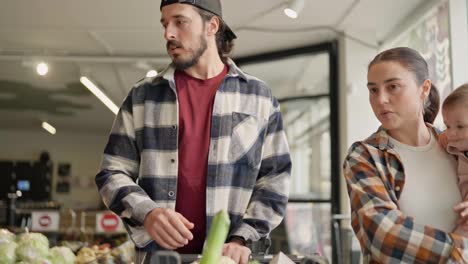 A-confident-brunette-man-with-a-beard-together-with-his-wife-and-a-small-child-with-a-cart-choose-products-and-do-their-shopping-in-the-supermarket