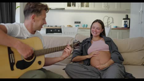 Happy-pregnant-brunette-woman-sitting-on-the-sofa-and-listening-to-her-husband-playing-guitar-at-home-on-the-weekend