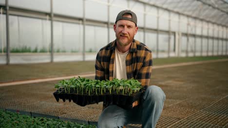 Portrait-of-a-happy-guy-farmer-in-a-cap-in-a-plaid-shirt-who-holds-seedlings-of-young-plants-in-his-hands-in-a-greenhouse-on-the-farm