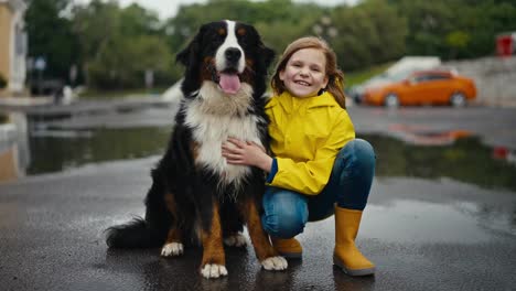 Portrait-of-a-happy-blonde-girl-in-a-yellow-jacket-who-sits-next-to-her-large-black-and-white-purebred-dog-and-in-the-park-after-the-rain