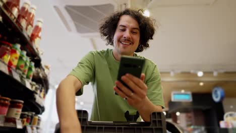 Happy-brunette-guy-with-curly-hair-watches-funny-videos-on-his-phone-while-shopping-and-driving-with-a-cart-in-the-supermarket