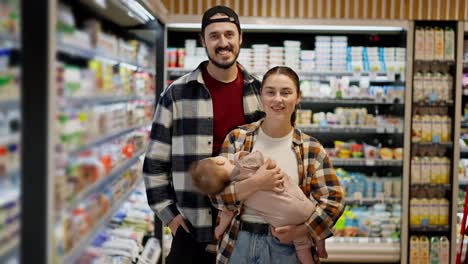 Portrait-of-a-happy-brunette-man-in-a-plaid-shirt-with-his-wife-and-little-child-near-dairy-products-in-a-supermarket.-Family-shopping-of-a-husband-and-wife-together-with-an-infant-in-a-supermarket