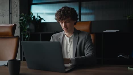 Un-Joven-Feliz-Con-Cabello-Rizado-Con-Un-Traje-Gris-Trabajando-Y-Escribiendo-En-Una-Computadora-Portátil-Gris-Mientras-Está-Sentado-En-Una-Mesa-En-Una-Oficina-Moderna