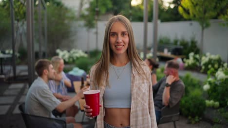 Retrato-De-Una-Chica-Rubia-Feliz-Con-Una-Camisa-Clara-Sosteniendo-Un-Vaso-Rojo-En-Sus-Manos-Y-Posando-Cerca-De-Su-Compañía-De-Vacaciones-En-El-Patio-De-Una-Casa-De-Campo.