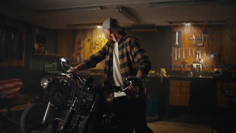 A-confident-guy-in-a-checkered-shirt-brings-in-a-motorcycle-and-prepares-for-repairs-in-the-workshop